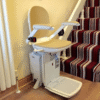A reconditioned Acorn or Brooks 180 Curved Stairlift with a beige chair, armrests, and footrest is installed at the bottom of a striped carpeted staircase in a home, ready for use.