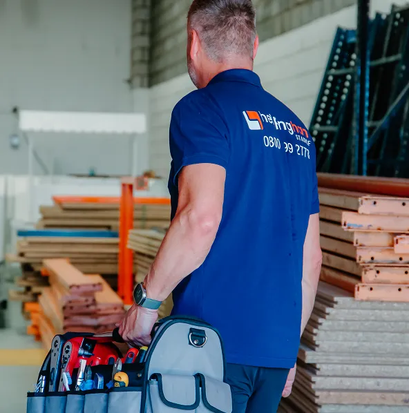 A man in a blue company-branded polo stands in a workshop holding a large tool bag. Behind him are wooden boards and metal shelves. He is installing the Brooks 140 Outdoor Stairlift - New, with his face turned away from the camera.