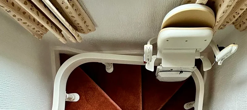 View from the top of a narrow, carpeted staircase with a stairlift installed along the bannister, winding down in a tight curve with white walls and beige curtains above.