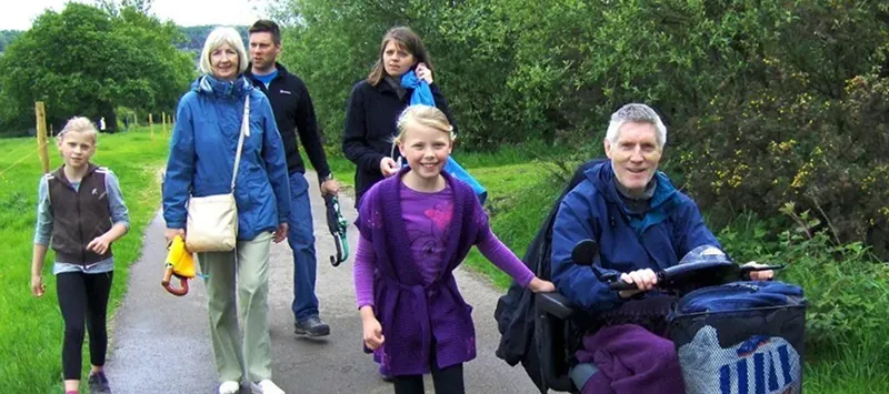 A group of six people, including children and adults, walk along a tree-lined path. One man uses a motorised wheelchair, and others walk beside him, smiling and enjoying the outdoors.